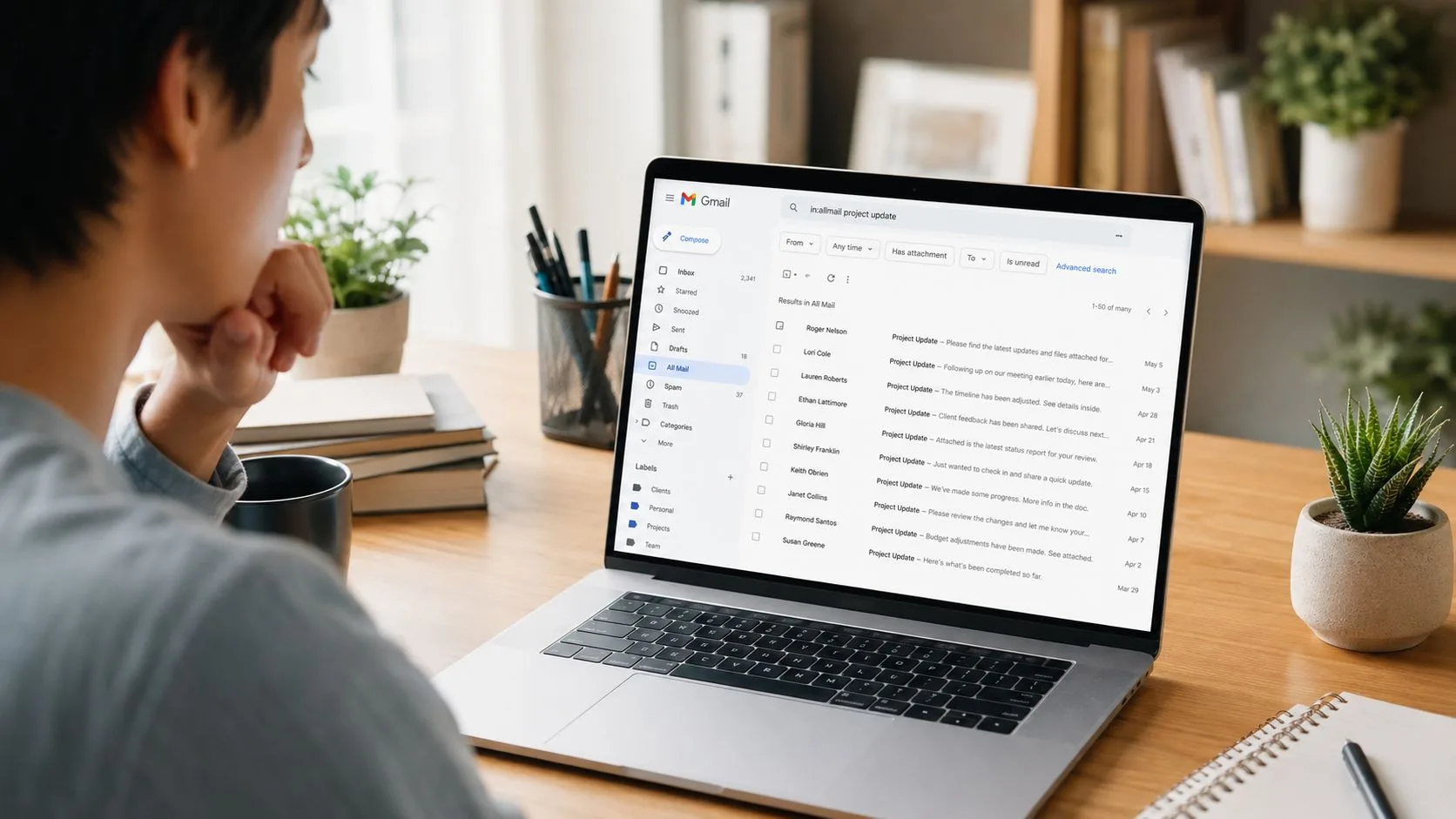 A young man sits at a wooden desk looking at a Gmail inbox screen on his laptop.